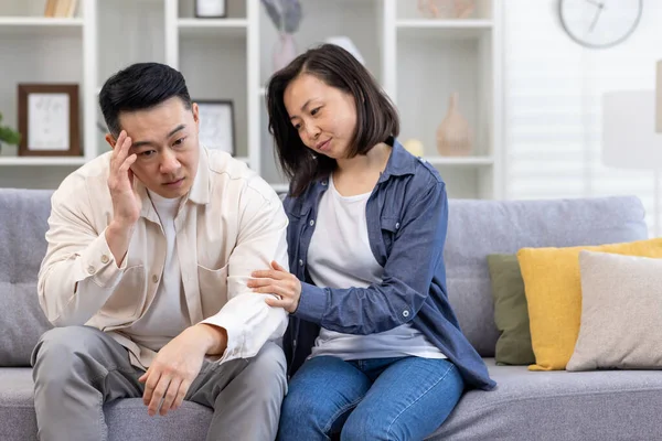 Family psychological support, Asian couple sitting at home on sofa thoughtful, woman comforting and supporting depressed man.