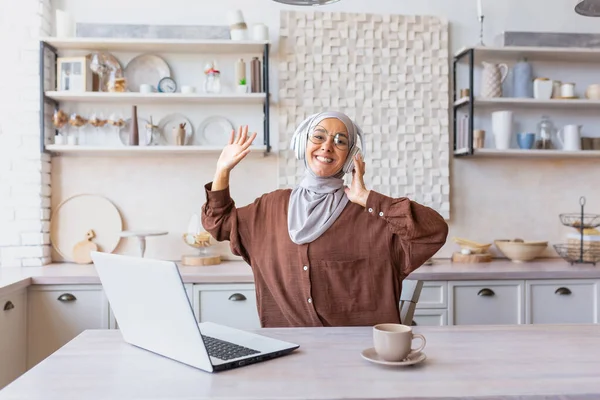 Genç Latin Amerikalı tesettürlü ve kulaklıklı bayan öğrenci evde laptopun önünde ders çalışıyor, eller yukarı, dans ediyor, konferans dinliyor. Gülümsüyor, kameraya bakıyor..