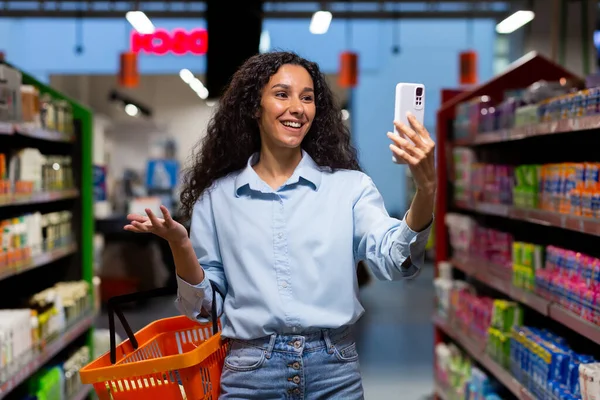 A young Latin American woman, blogger stands in a supermarket with a basket among the shelves with goods and talks on a video call from the phone, consults, asks what to buy, smiles.