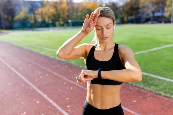 stock image Tired and upset mature female athlete checking heart rate on smart watch fitness bracelet, runner in stadium after active exercise and jogging on sunny day, blonde woman in sportswear.