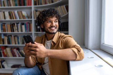 Genç başarılı İspanyol öğrencinin yakın portresi, pencereden bakan kıvırcık saçlı adam gülümseyen ve mutlu, üniversitede akademik kütüphanede otururken telefonu elinde tutan genç..
