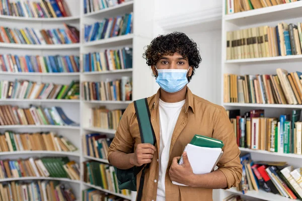 Portrait of a young male student inside a library studying at a university, hispanic man with curly hair looking at the camera holding books in his hands, the guy is wearing a protective face mask.