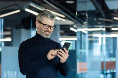 Ofisin içinde yetişkin bir adam pencerenin yanında duruyor, iş adamı telefonda mesaj yazıyor ve internet sayfalarını karıştırıyor, gri saçlı patron internette haberleri okuyor..