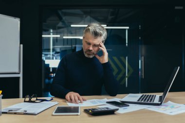 Ofis içinde belgelerle çalışan ciddi bir finansör iş analisti, ofiste hesap makinesi ve dizüstü bilgisayarla işlem yapan yetişkin bir muhasebeci..