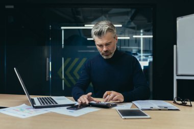 Ofis içinde belgelerle çalışan ciddi bir finansör iş analisti, ofiste hesap makinesi ve dizüstü bilgisayarla işlem yapan yetişkin bir muhasebeci..