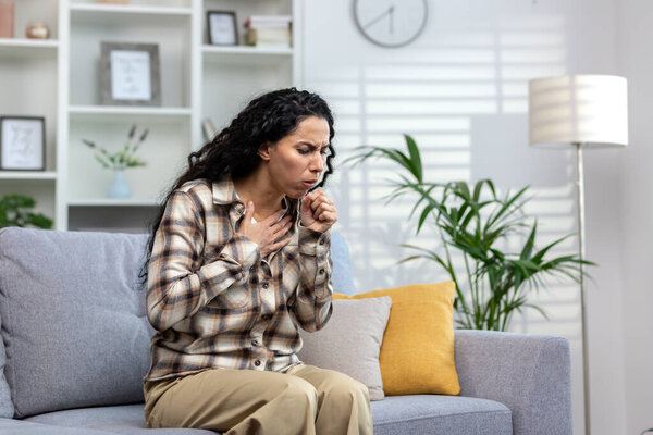 Woman having cold and sick coughing sitting on couch at home, hispanic woman in living room with cold and flu.