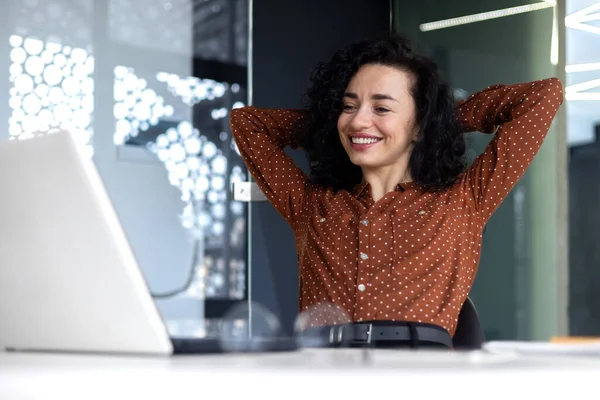 Happy thinking woman at workplace inside office, business woman with hands behind head completed work well, latin american woman satisfied with achievement results looking at laptop screen.