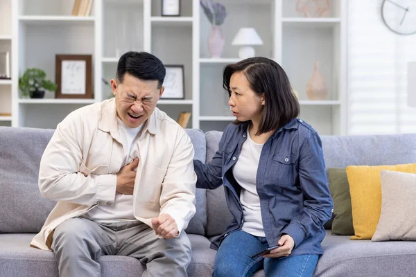 Young Asian family sitting on sofa at home. A man holds his hand to his chest, heart attack, stroke, panic attack, feels severe pain. Shocked woman tries to help, give first aid.