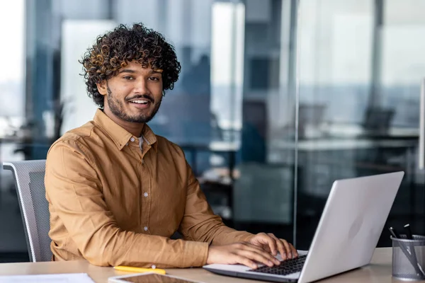 Portrait of young successful Indian man at workplace inside office, businessman smiling and looking at camera, man at work using laptop, programmer with curly hair coding software.