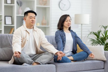 Evdeki mutlu Asyalı aile kanepede meditasyon yapıyor, çift erkek ve kadın oturma odasında lotus pozisyonunda oturuyor ve gözleri kapalı dinleniyor..
