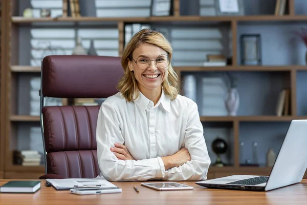 stock image Portrait of an older beautiful blonde woman, director, boss, founder of the company in glasses and a white shirt. Sitting at the desk in the office, working on a laptop and smiling at the camera.