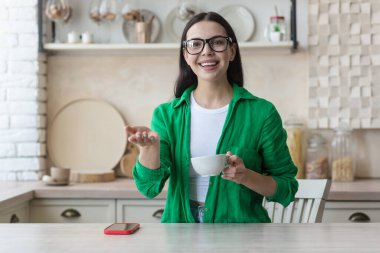 Genç bir kadın mutfakta elinde bir fincan kahveyle kameranın önünde oturuyor ve video kaydediyor, blog kaydediyor, web sitesi, büyüme ve gelişme eğitimi veriyor, kamerayı işaret ediyor, gülümsüyor..