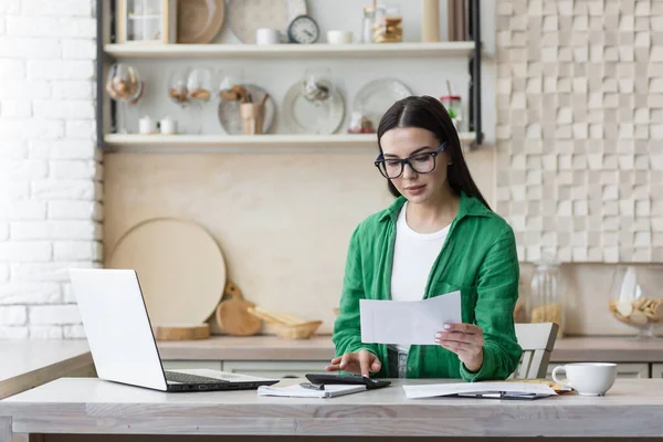 stock image Work remotely. A young beautiful businesswoman works from home. Sitting at home in kitchen working with laptop, documents and online calculator. Dressed in glasses and a green shirt.