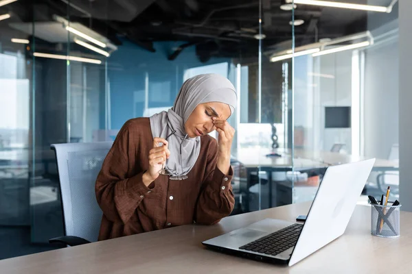 Tired and overworked business woman in hijab at workplace, muslim woman massaging eyes with glasses off, female worker working late on project.