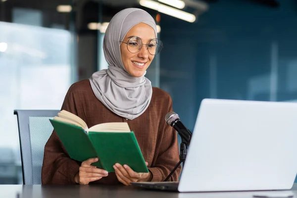 Young beautiful muslim woman in hijab working in modern office using professional microphone to record audio podcast reading religious book online radio.