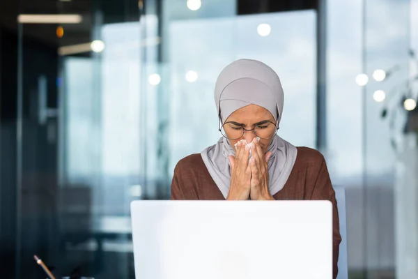 Businesswoman sick at workplace, Muslim woman in hijab sneezing and having a runny nose, female worker inside office wiping nose with tissue, working with laptop.