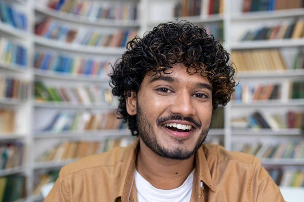 Close-up portrait of young hispanic student, man with curly hair smiling and looking at camera, young scholar studying inside academic library.