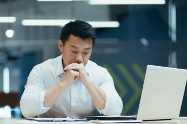 Masadaki ofiste oturan üzgün Asyalı erkek işadamı, düşünceli bir şekilde başını ellerine yaslıyor. İşteki mali sorunlar, iflas, işten çıkarma.
