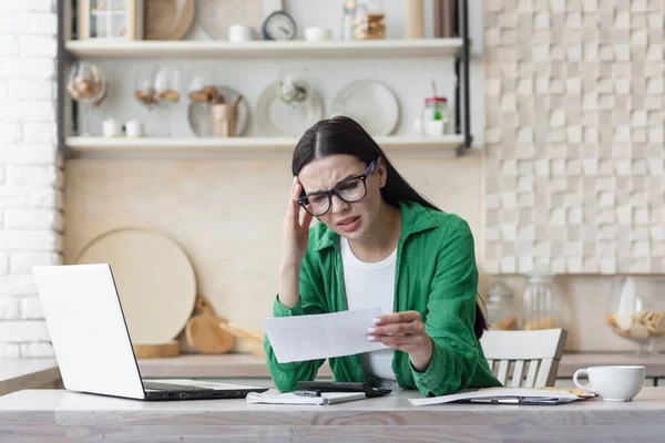 stock image Worried young woman reading a received letter while sitting in the kitchen at home.