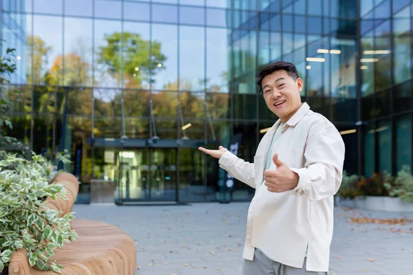 A young Asian man, a businessman, a freelancer, is standing outside an office, a skyscraper, and points to the entrance with his hands. invites with a gesture to come inside, see his project, work.