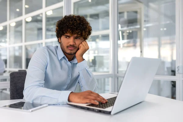 Upset young businessman working inside office with laptop, man fails to complete technical project on time, hispanic man frustrated sitting at desk.