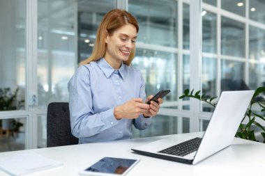 Başarılı bir iş kadını patronu modern parlak ofisin içindeki iş yerinde telefonu kullanıyor, ofis çalışanı akıllı telefondan gülümseyen internet mesajı yazıyor.