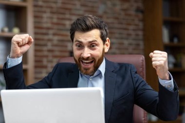 Başarılı bir iş adamı zaferi ve başarıyı kutluyor. Yetişkin yetişkin erkek patron elde ettiği başarılardan memnun. Zafer jestleri yapan finansörleri iş yerinde dizüstü bilgisayar kullanarak mutlu ediyor.