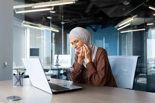 stock image Toothache at the workplace. A young woman working in the office at a table behind a laptop holds her cheek, feels severe toothache, needs medical help from a dentist.