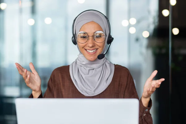 Close-up photo. Online teaching. Portrait of young Arab woman in hijab and headset, teacher sitting in front of laptop, explaining, talking, gesturing with hands. Conducts distance education.