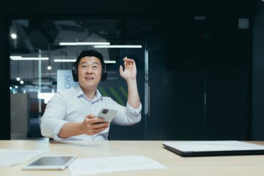 Dinlen, işe ara ver. Kulaklıklı genç bir Asyalı ofis masasında oturuyor, telefonu elinde tutuyor ve müzik dinliyor, dans ediyor, rahatlıyor..