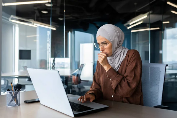 Illness at work. A young Arab woman in a hijab sits in the office at a desk and coughs. He covers his mouth with his hand, feels bad.