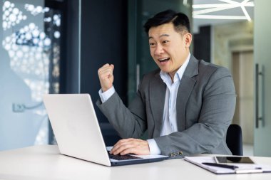 Başarılı Asyalı yatırımcı zaferi ve zaferi kutluyor, takım elbiseli olgun patron ofis içinde dizüstü bilgisayarla çalışıyor, adam online galibiyet haberi aldı.