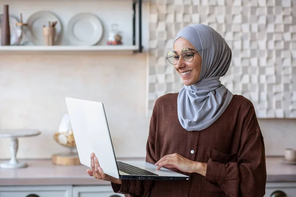 Joyful and successful business woman in hijab at home using laptop while standing, muslim woman working remotely.