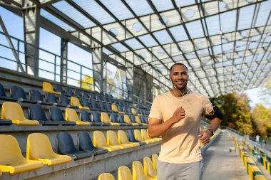 Genç adam stadyumda spor yapıyor, sabah kulaklıkla koşuyor, telefonla koşuyor, maratona hazırlanıyor, kameraya gülümsüyor..