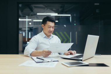 Ofis işleri. Genç Asyalı bir adam masada otururken dizüstü bilgisayar ve belgeler üzerinde çalışıyor..
