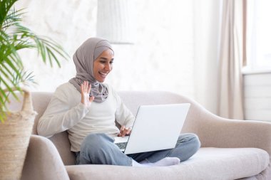 Çevrimiçi iletişim. Genç, tesettürlü Müslüman bir kadın evinde, nilüfer çiçekli koltukta oturuyor dizüstü bilgisayarını kucağında tutuyor ve bir video görüşmesinde konuşuyor..