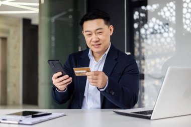 İş yerinde bilgisayarla çalışan başarılı Asyalı patron, telefon ve banka kartı tutan işadamı, rezervasyon ve online para transferi için uygulama kullanan adam..