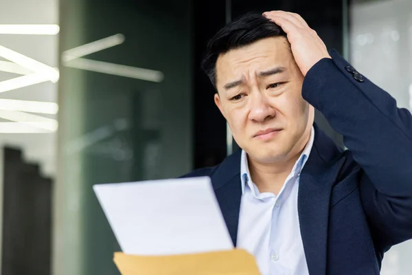 Frustrated and sad depressed businessman received mail envelope notification bad news, asian man at workplace inside office working with laptop, unhappy with achievement results.