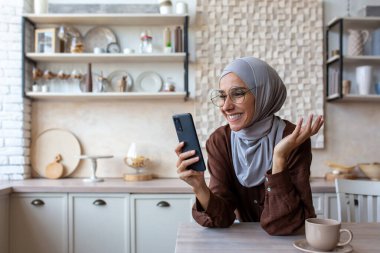 Yakın plan fotoğraf. Gülümseyen tesettürlü, tesettürlü genç bir kadın mutfakta duruyor, masaya yaslanıyor ve cep telefonuyla konuşuyor..