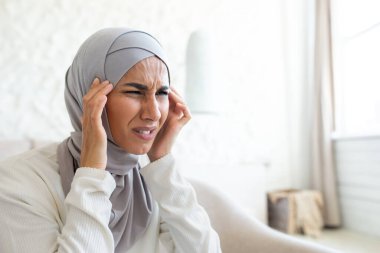 Yakın plan fotoğraf. Bunalımlı ve yorgun. Tesettürlü genç bir kadının portresi elleriyle başını tutuyor, şiddetli bir acı hissediyor, kendine masaj yapıyor, gerilimden buruşmuş..