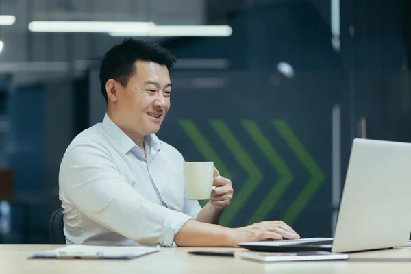 Asian smiling young man working in office on laptop, holding cup in hand, coffee break.