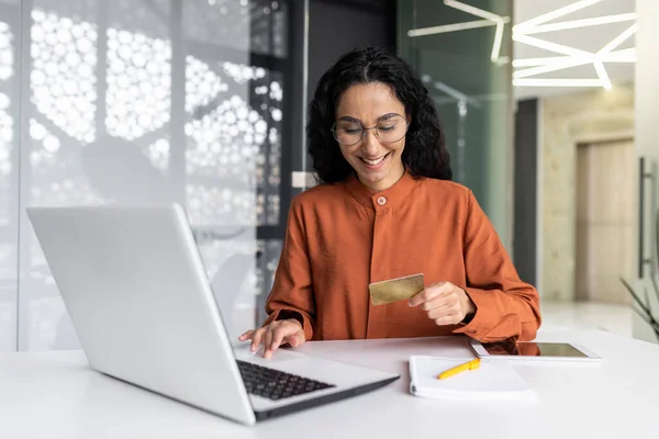 Young successful hispanic business woman working inside office at workplace, joyful female worker making online purchase in online store and booking services, holding bank credit card, using laptop.