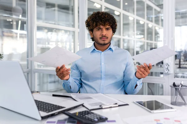 Portrait of angry disappointed financier inside office at workplace, hispanic businessman holding contracts reports in hands and looking nervously at camera, upset man behind paper work.