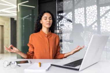 Young beautiful Hispanic web developer working inside office building, woman resting meditating at workplace with eyes closed, female worker using laptop to write code.