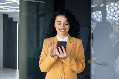 Telefonla gülümseyen başarılı İspanyol kadının, iş yerindeki iş kadınının elinde akıllı telefon okuyup sarı ceketli pencerenin yanında duran haberlerin yakın plan fotoğrafı..