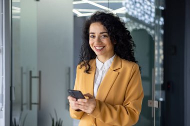 Ofiste başarılı bir iş kadını, sarı ceketli ve pencerenin yanında duran gözlüklü kadın patron portresi kameraya bakıyor ve gülümseyen küçük işletme sahibi telefon tutuyor, mesaj yazıyor.