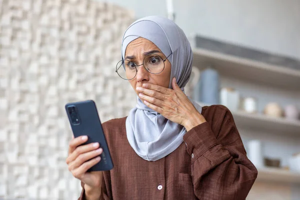 Young arab woman in hijab looking at phone in shock at home in kitchen. She covered her mouth with her hand in surprise, she received bad news.