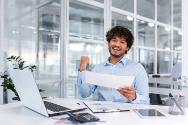 Portrait of successful hispanic businessman, man in shirt reading report and happy celebrating success satisfied with result holding hand up, worker inside office with laptop working on paper work.