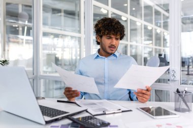 Pensive serious businessman reading financial report, hispanic businessman holding document in hands looking disappointed, working inside modern office with laptop behind paper work.