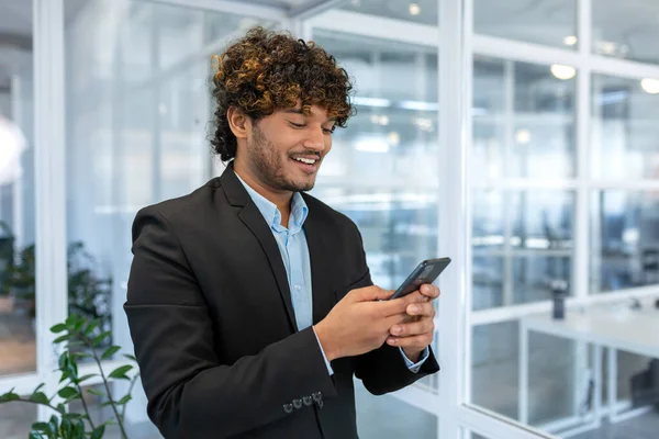 stock image Successful financier investor works inside office at work, businessman in business suit uses telephone near window, man smiles and reads good news online from smartphone.